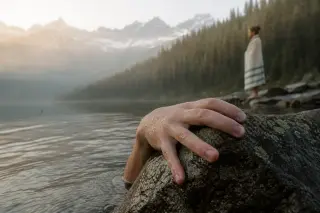 Swimmer's hand on wet stone at misty alpine lake, background shows forested slope and distant peaks in soft sunrise light, candid and atmospheric.