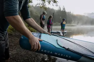 Close-up of hands steadying a paddleboard on an alpine lake shore at sunrise, with a family preparing in the background and pine forest reflections in the misty water.