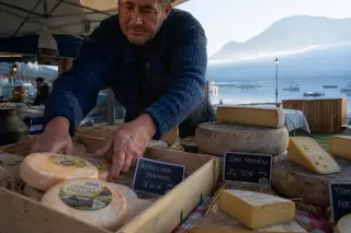 Local cheesemonger arranging fresh cheese at a lakeside market with misty mountains and water in the background on an early morning at Lake Annecy