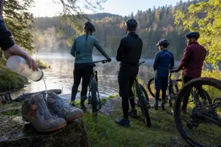 Cyclists pausing by Lake Longemer, cleaning shoes in dewy grass with misty forest and morning light in the background