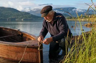 Elderly man tying knots beside a wooden dinghy at sunrise on a French alpine lake, with soft light and natural lakeshore details.