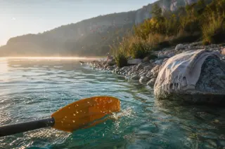 Paddle blade slicing through sunlit water of Lac de Chalain near a wild shoreline with cliffs and grasses, towel on stones in background, late afternoon light.