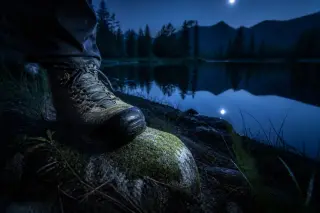 Boot stepping onto a mossy rock near a forested alpine lake at night, illuminated by a headlamp with lake and pine silhouettes in the background.
