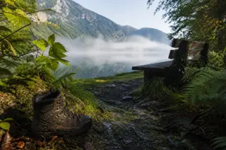 A single hiking boot beside a rustic bench on a misty lakeside path, Lac d'Aiguebelette and forested hills in soft morning light.