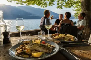 Freshly served fish dishes and wine on a rustic lakeside terrace, with relaxed diners and alpine lake view in warm afternoon light