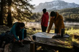 Hikers consulting a map and adjusting gear at a mossy table by a misty alpine lake in Haute-Savoie at sunrise, with snow-capped mountains in the background.