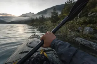 Inside a kayak at dawn on Lac de Serre-Ponçon, hands paddling past misty pine shore, droplets and natural light creating a serene, immersive atmosphere