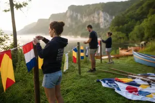 Volunteers preparing rowing boats and tying festival flags on a misty jurassic lakeshore at dawn, hands and details in focus, natural and authentic ambiance.