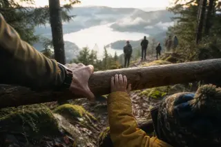 Parent and child hands on a wooden rail overlooking a misty lake with hikers and pine forest in warm sunlight.