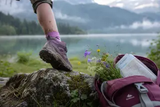 Close-up of a child’s muddy hiking shoe stepping on a mossy rock near Lake Annecy, with wildflowers and a map in a backpack, Alpine forest and misty lake in the background under soft morning light.